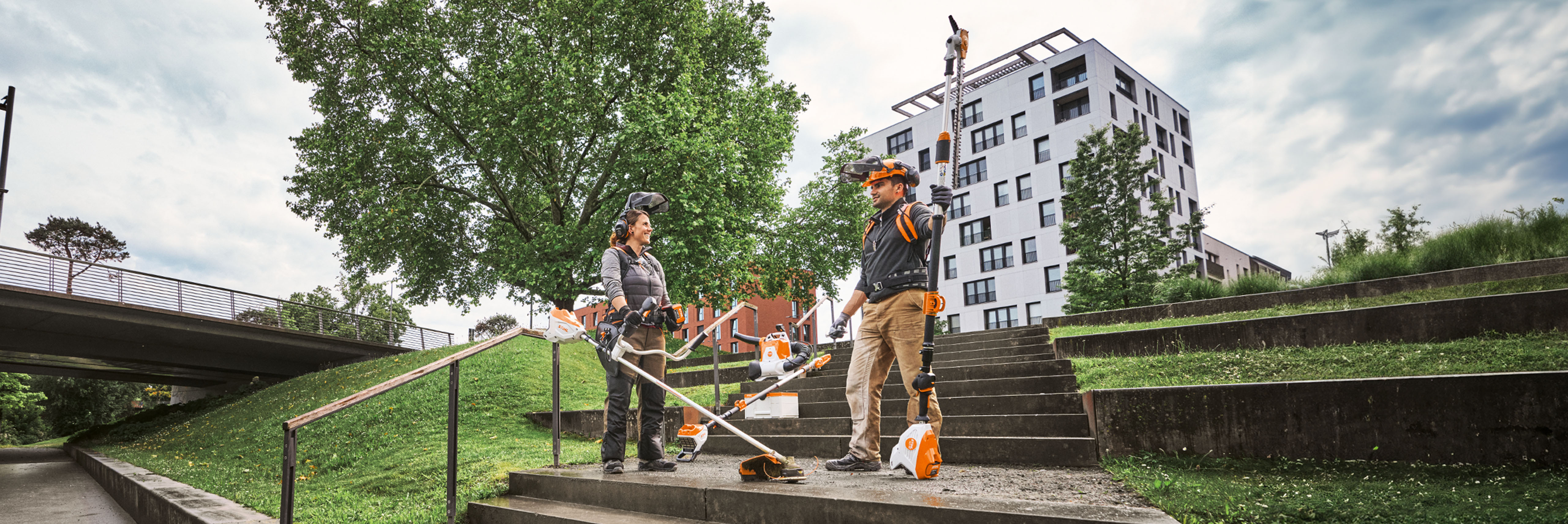 Dwóch profesjonalistów zajmujących się kształtowaniem krajobrazu używa narzędzi STIHL Pro i sprzętu akumulatorowego na miejskim terenie zielonym ze schodami i drzewami.