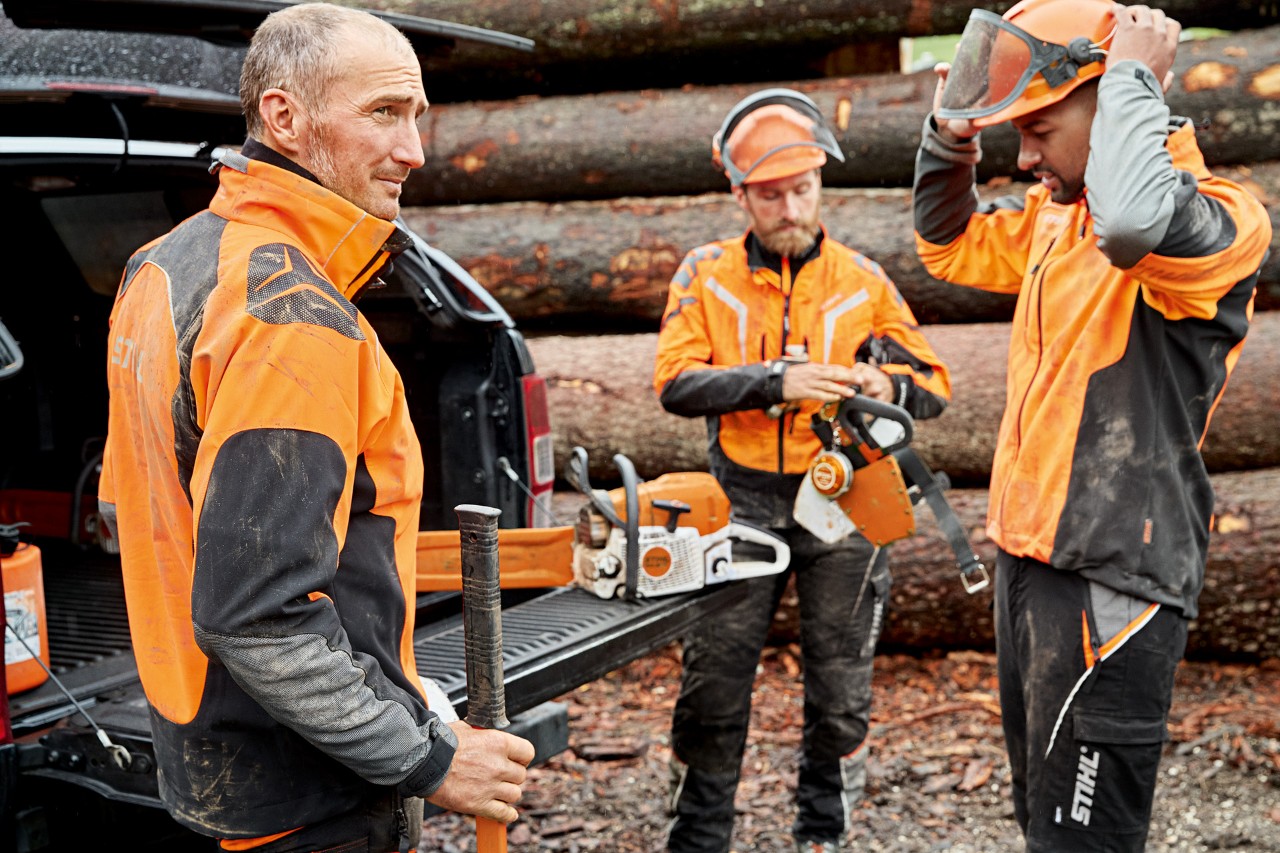 Trzej mężczyźni w odzieży ochronnej STIHL przed stosem drewna przygotowują się do pracy pilarką STIHL