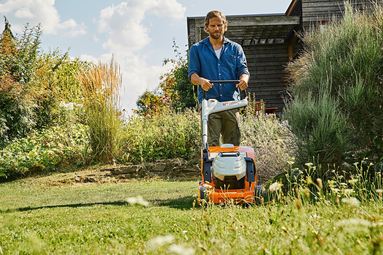 Mężczyzna jest w trakcie koszenia zaniedbanej i zarośniętej trawy akumulatorową kosiarką STIHL RMA 448 PC w ogrodzie otoczonym wysokimi krzewami i roślinami